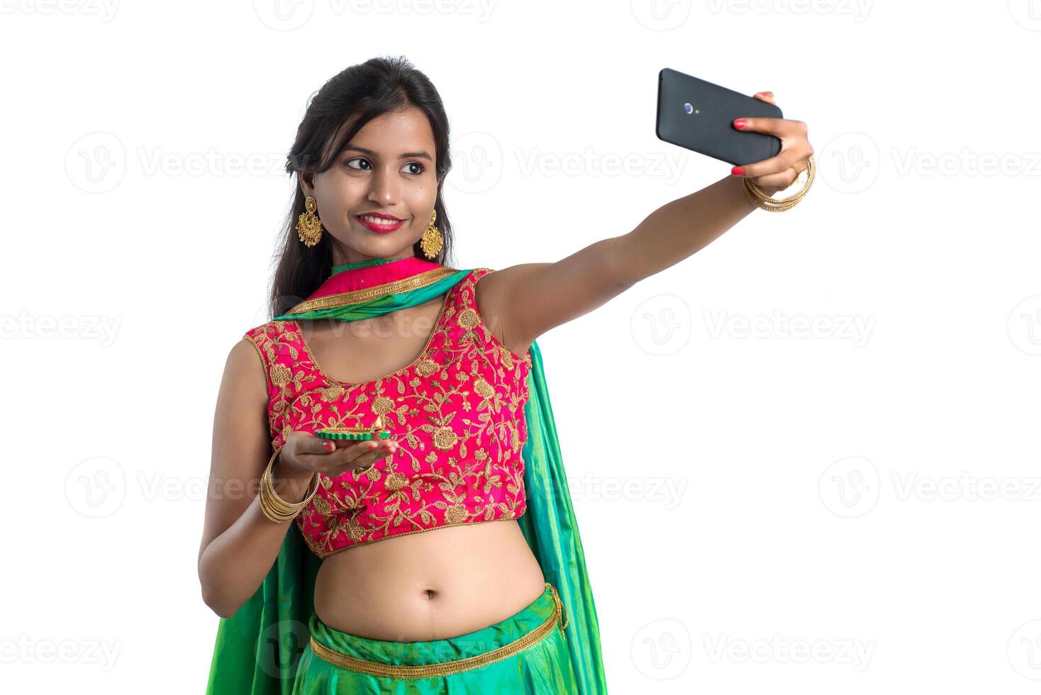 Beautiful young happy girl taking a selfie with clay lamp or diya during the festival of light Diwali using a smartphone on a white background photo