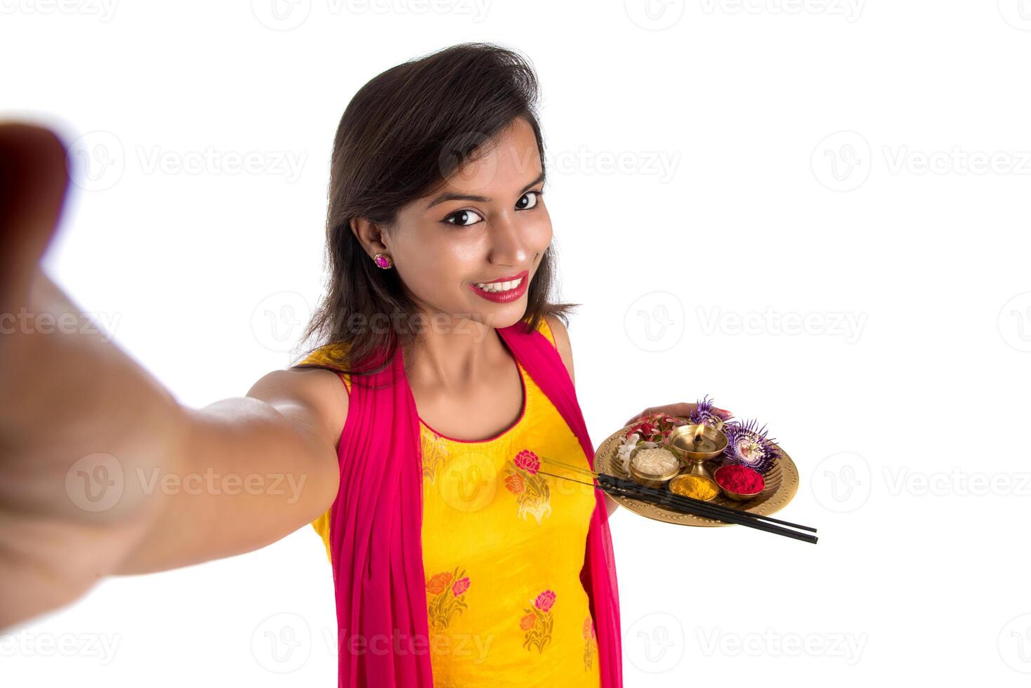 Beautiful young happy girl taking selfie with pooja thali using a mobile phone or smartphone on a white background photo