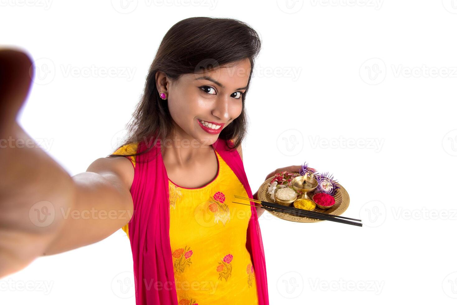 Beautiful young happy girl taking selfie with pooja thali using a mobile phone or smartphone on a white background photo