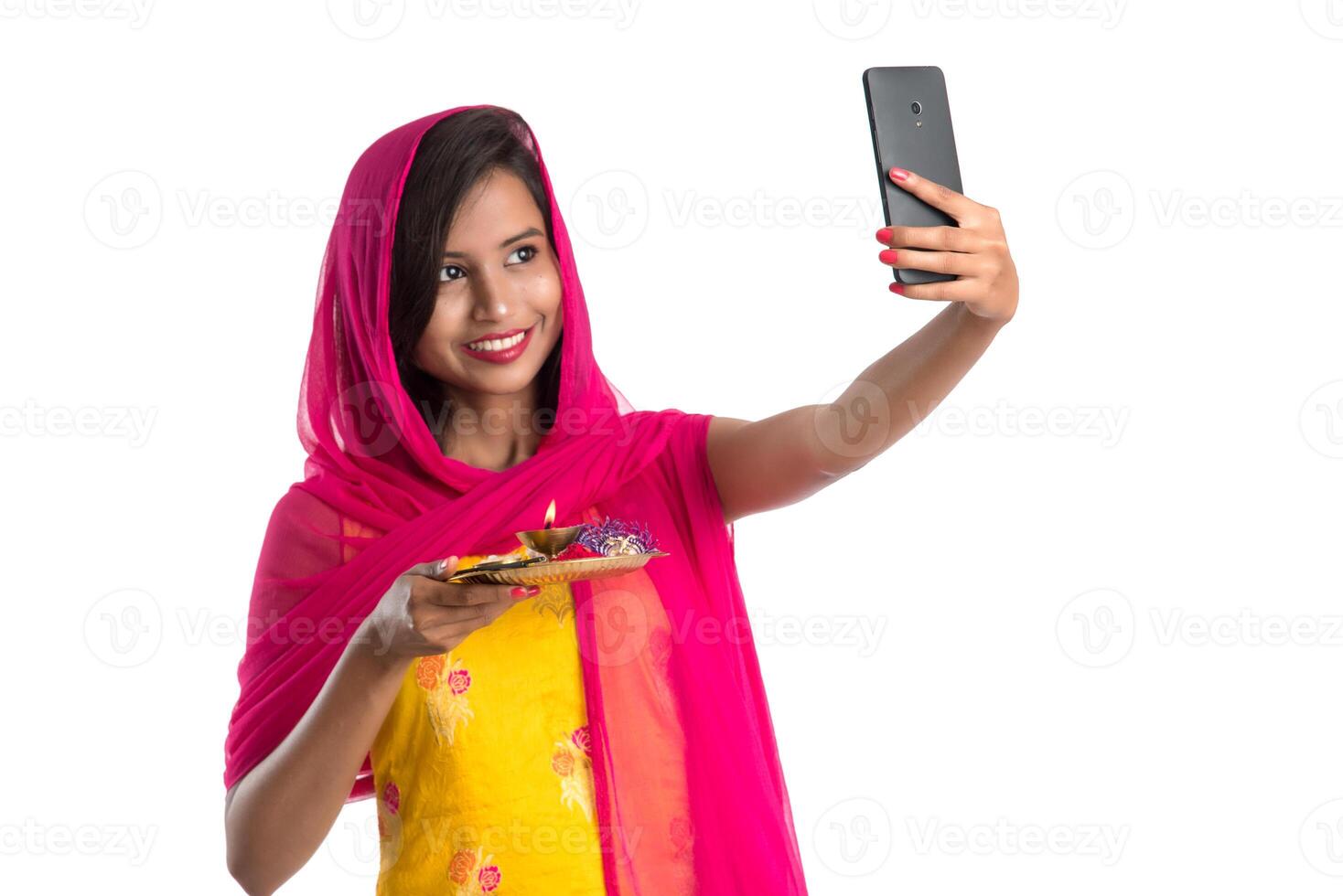 Beautiful young happy girl taking selfie with pooja thali using a mobile phone or smartphone on a white background photo
