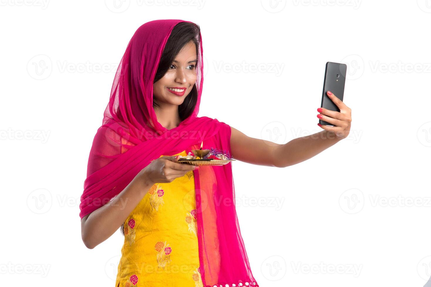 Beautiful young happy girl taking selfie with pooja thali using a mobile phone or smartphone on a white background photo