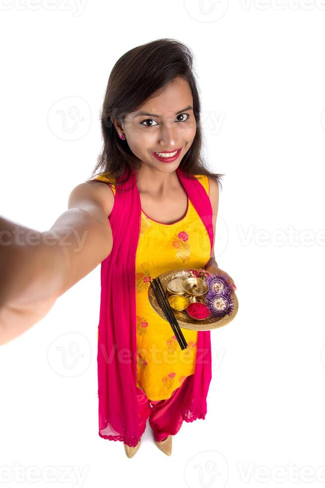 Beautiful young happy girl taking selfie with pooja thali using a mobile phone or smartphone on a white background photo