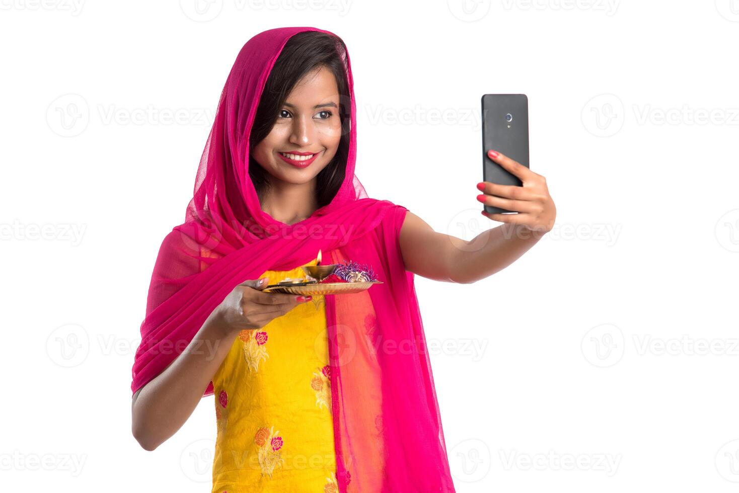 Beautiful young happy girl taking selfie with pooja thali using a mobile phone or smartphone on a white background photo
