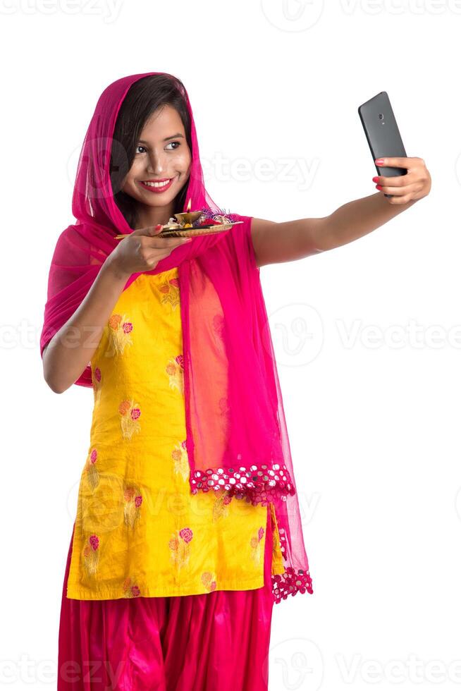 Beautiful young happy girl taking selfie with pooja thali using a mobile phone or smartphone on a white background photo