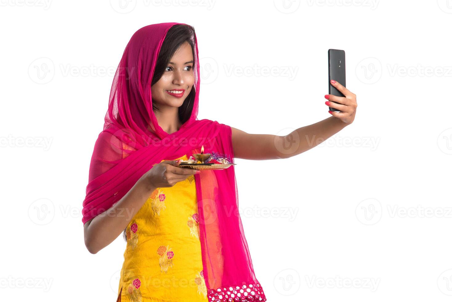 Beautiful young happy girl taking selfie with pooja thali using a mobile phone or smartphone on a white background photo