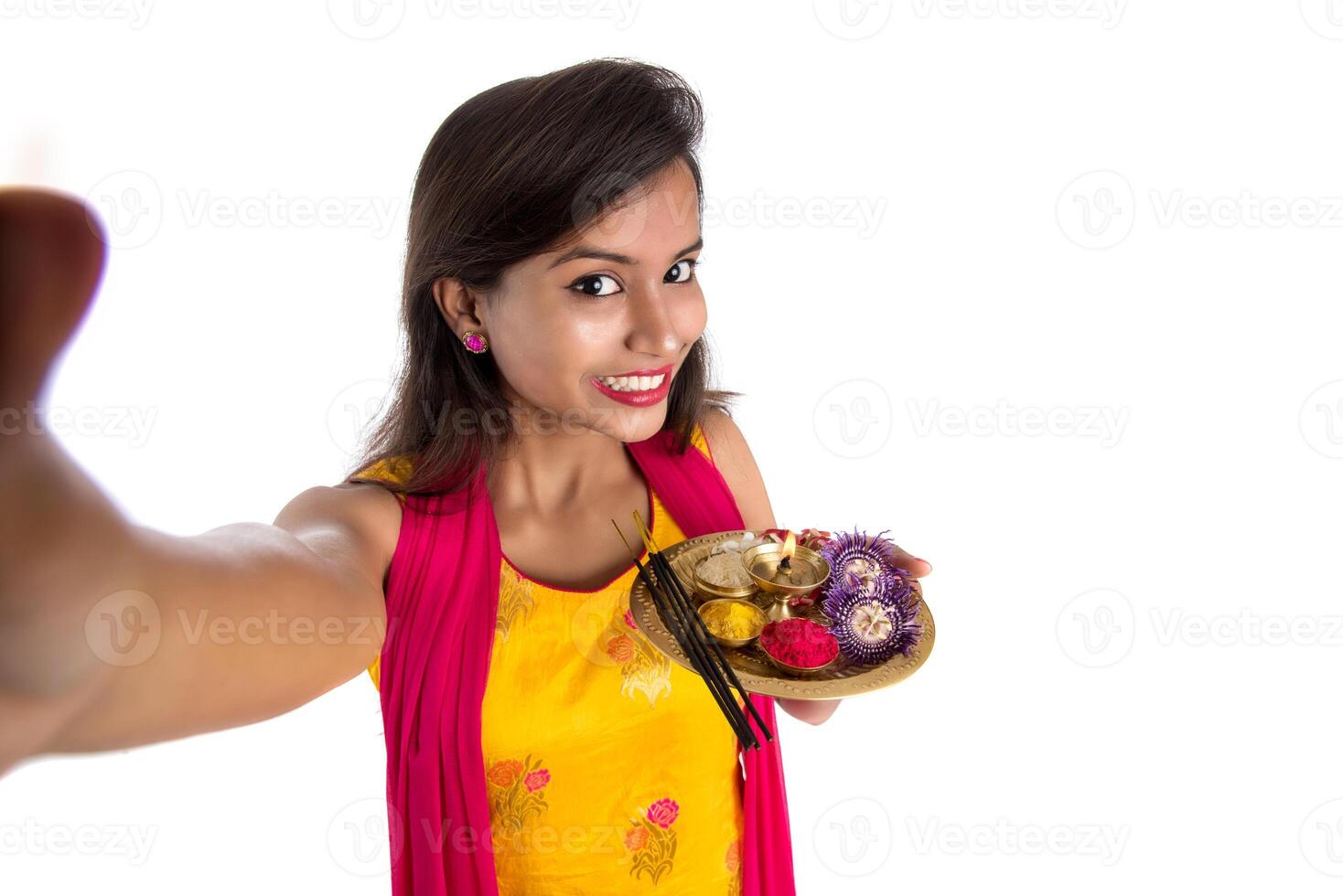 Beautiful young happy girl taking selfie with pooja thali using a mobile phone or smartphone on a white background photo