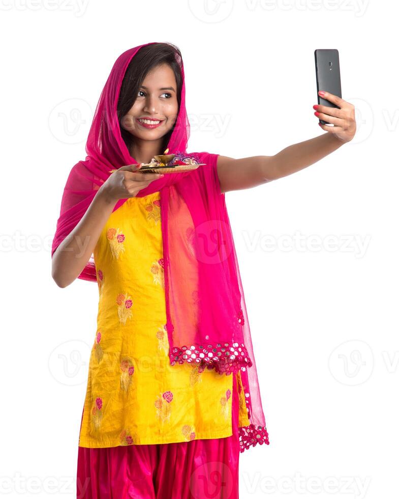 Beautiful young happy girl taking selfie with pooja thali using a mobile phone or smartphone on a white background photo