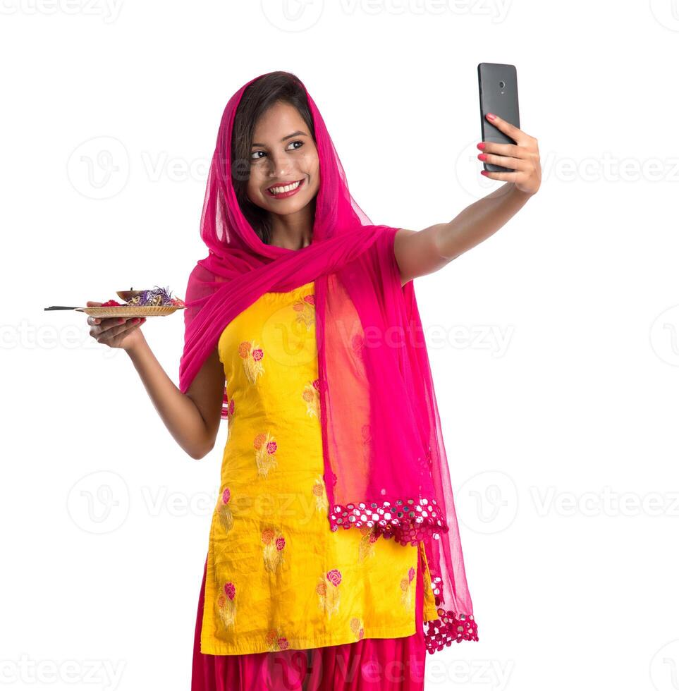 Beautiful young happy girl taking selfie with pooja thali using a mobile phone or smartphone on a white background photo