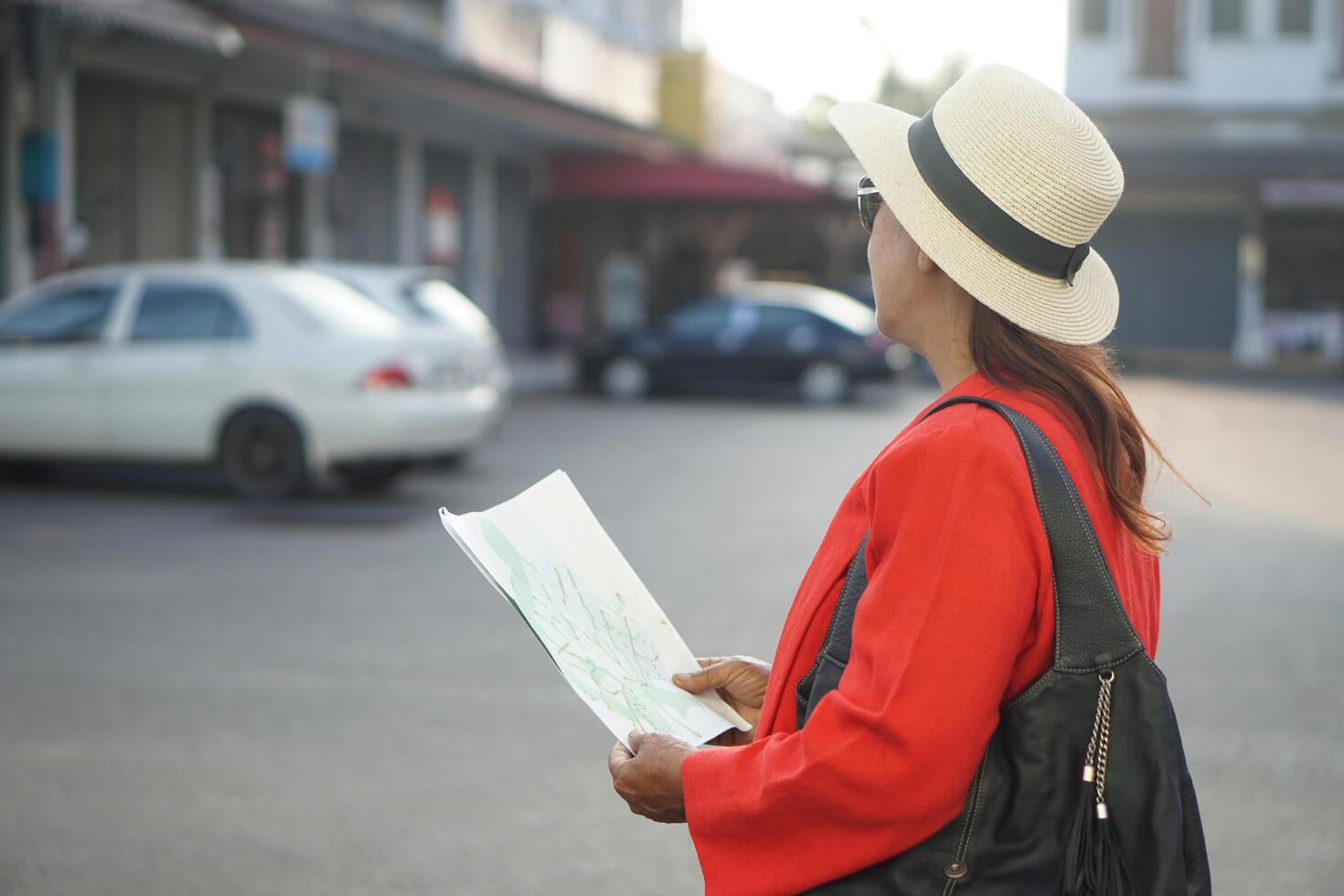 Asian woman tourist is standing outdoor, hold paper map during travel alone. Senior woman spends time traveling after retirement on vacation. Concept, travelling lifestyle. photo