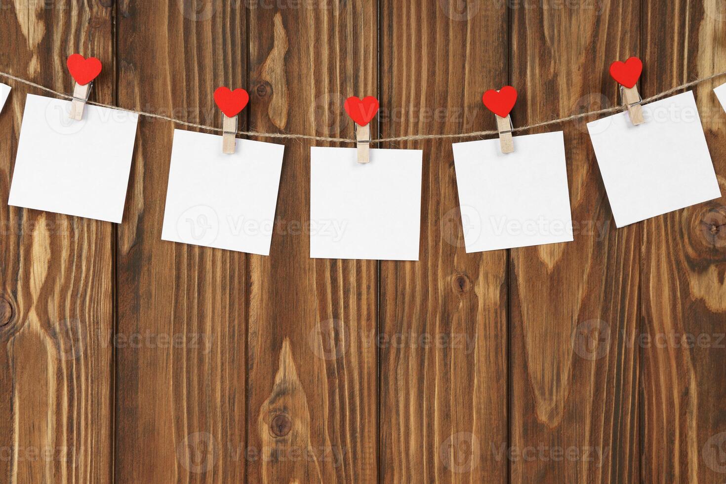 Clothespins with red hearts and empty sheets of paper on string against brown wooden background photo