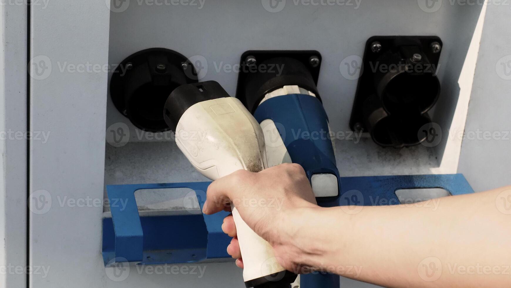 Driver of electric car hold batteries charger plug. Hand holding EV charging plug in front of camera with blurred background of outdoor. Sustainable energy powered electric charging station background photo