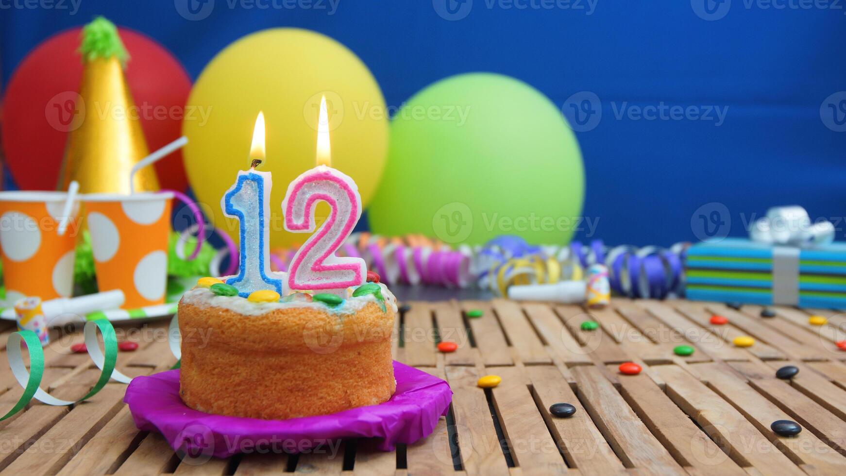 Birthday cake 12 with candles on rustic wooden table with background of colorful balloons, gifts, plastic cups and candies with blue wall in the background. Focus is on cake photo