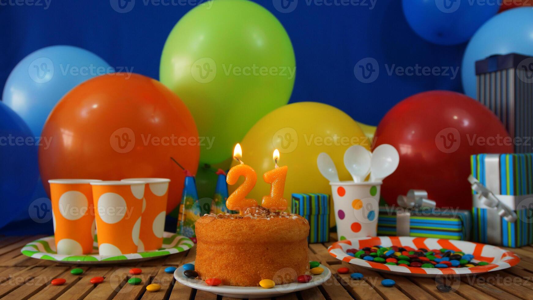 Birthday cake 21 on rustic wooden table with background of colorful balloons, gifts, plastic cups and plastic plate with candies and blue wall in the background photo