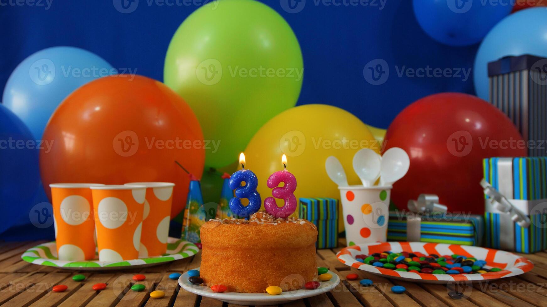 Birthday cake 33 on rustic wooden table with background of colorful balloons, gifts, plastic cups and plastic plate with candies and blue wall in the background photo