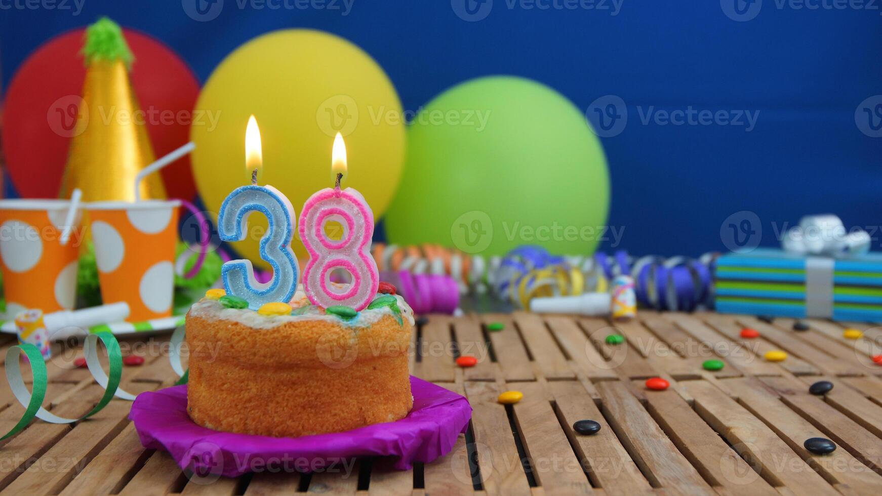 Birthday cake 38 with candles on rustic wooden table with background of colorful balloons, gifts, plastic cups and candies with blue wall in the background. Focus is on cake photo