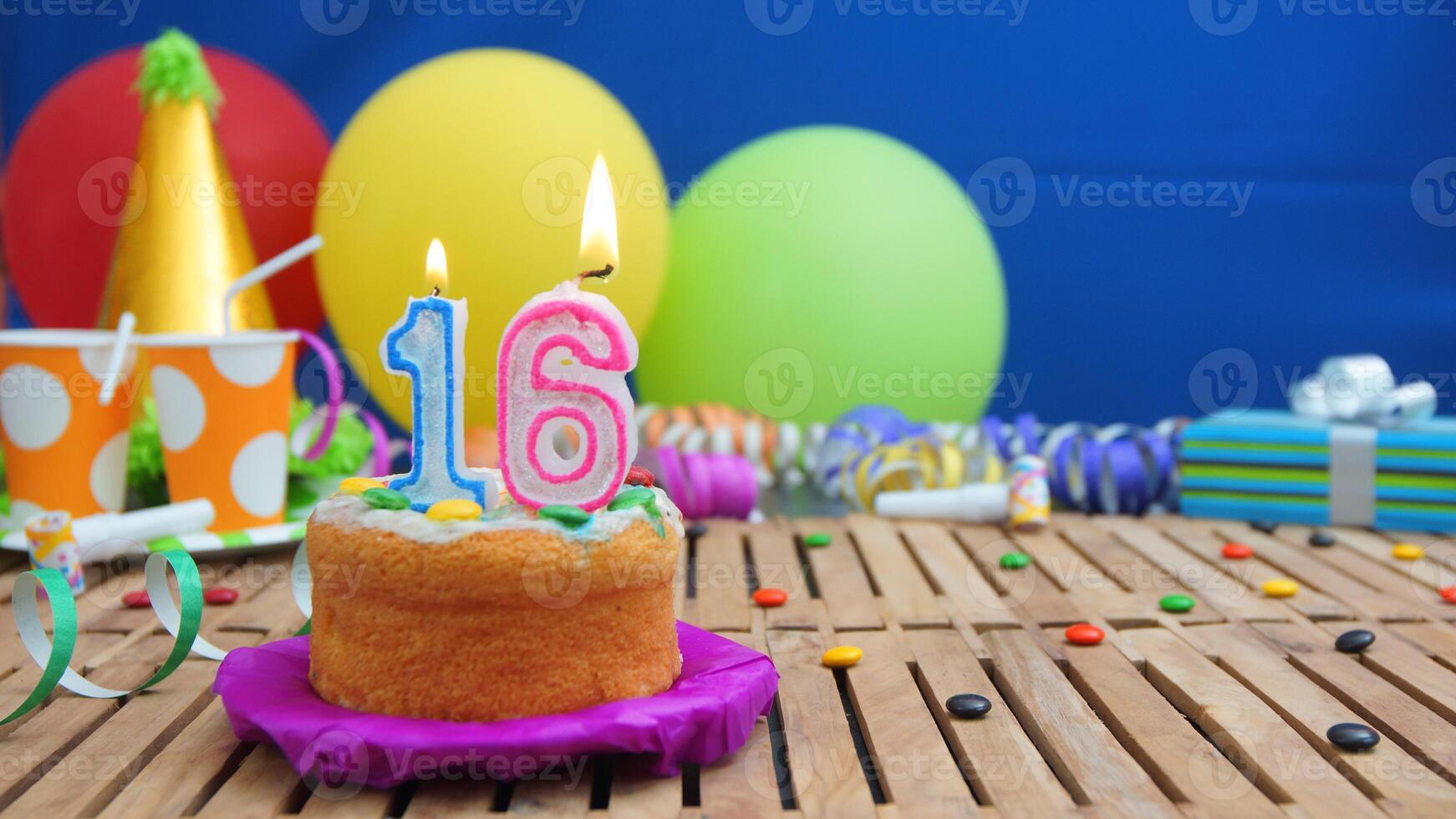 Birthday cake 16 with candles on rustic wooden table with background of colorful balloons, gifts, plastic cups and candies with blue wall in the background. Focus is on cake photo