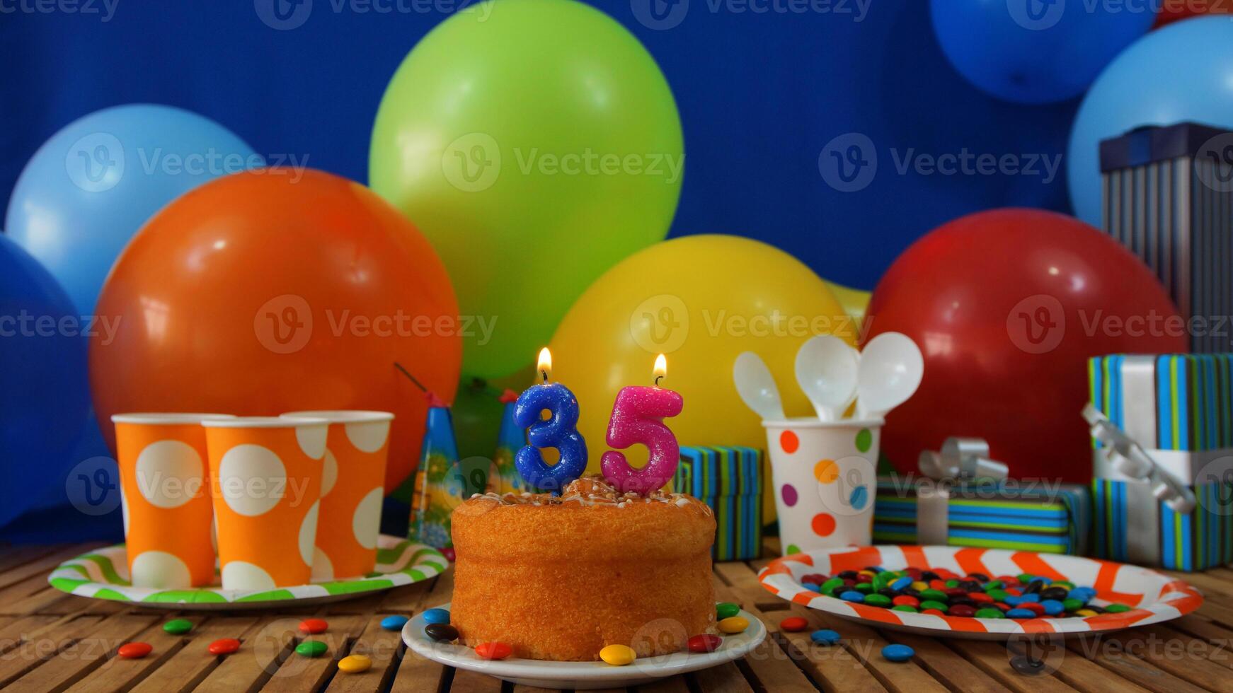 Birthday cake 35 on rustic wooden table with background of colorful balloons, gifts, plastic cups and plastic plate with candies and blue wall in the background photo