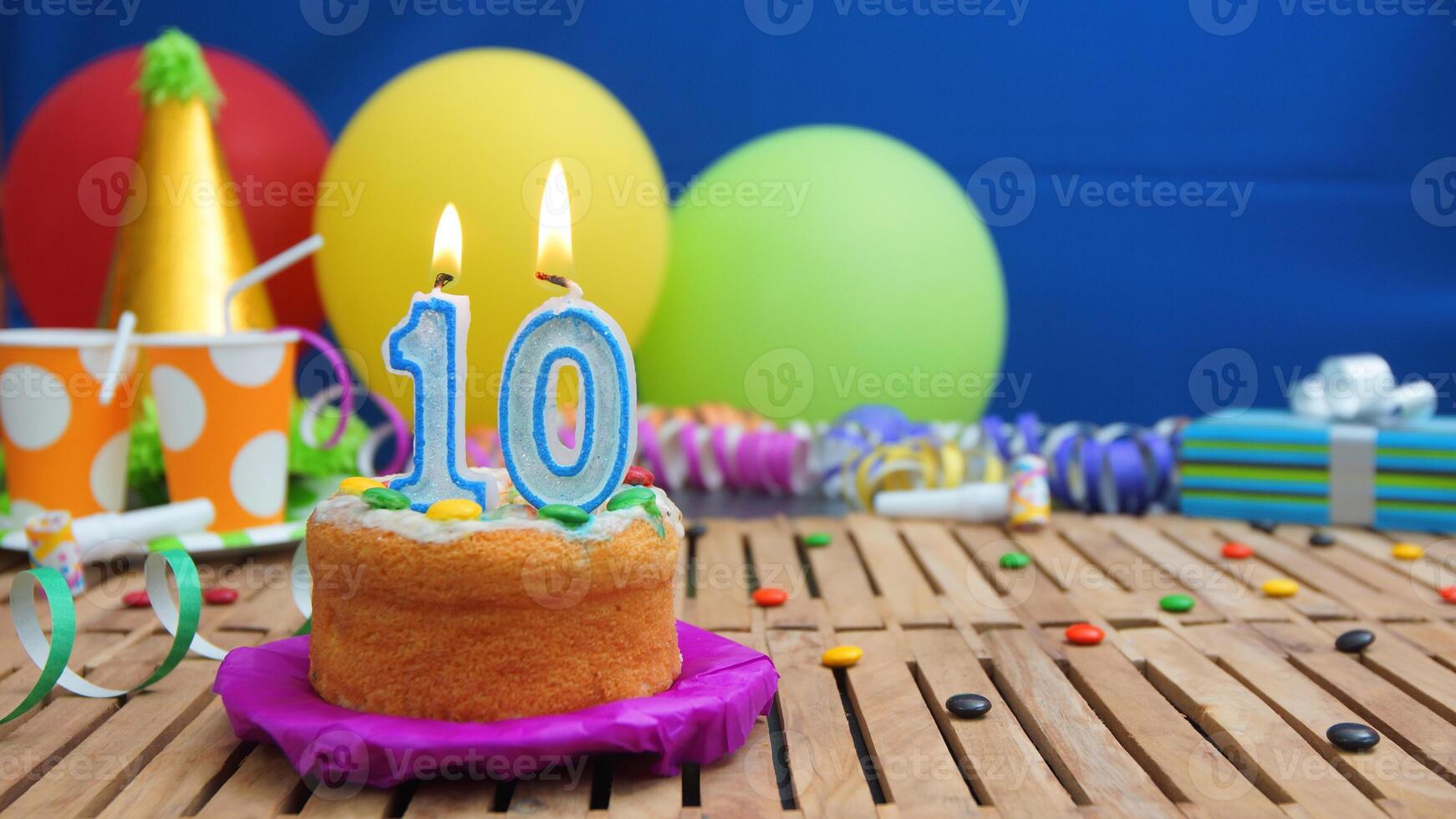 Birthday cake 10 with candles on rustic wooden table with background of colorful balloons, gifts, plastic cups and candies with blue wall in the background. Focus is on cake photo