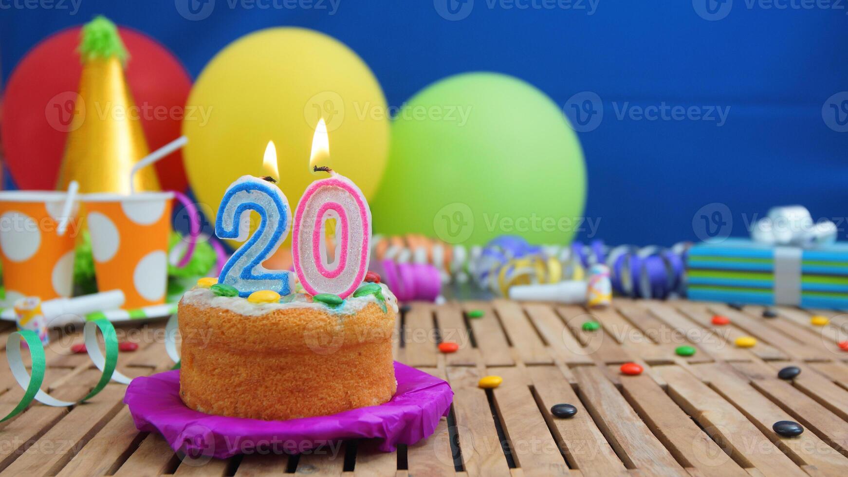 Birthday cake 20 with candles on rustic wooden table with background of colorful balloons, gifts, plastic cups and candies with blue wall in the background. Focus is on cake photo