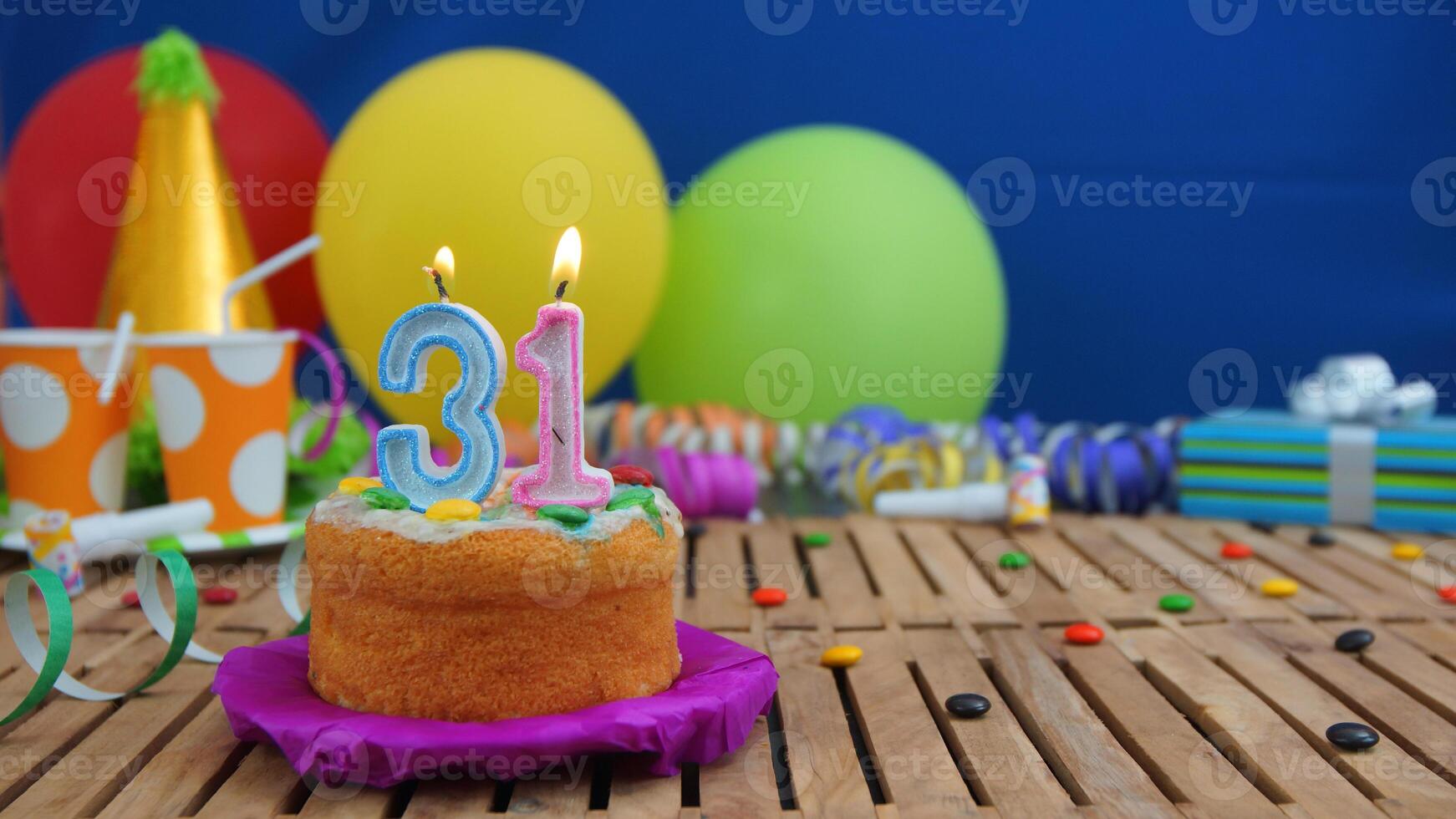 Birthday cake 31 with candles on rustic wooden table with background of colorful balloons, gifts, plastic cups and candies with blue wall in the background. Focus is on cake photo