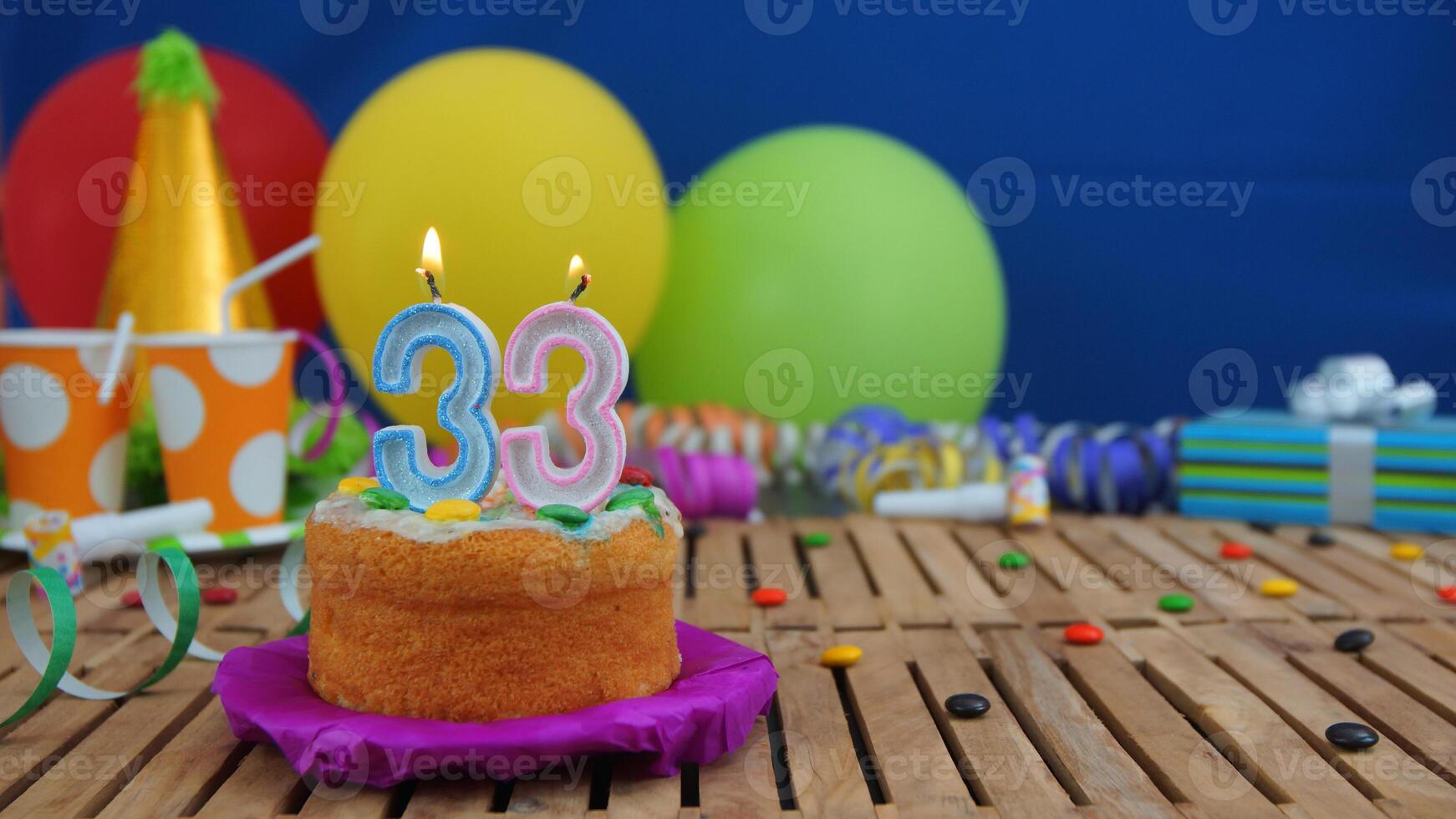 Birthday cake 33 with candles on rustic wooden table with background of colorful balloons, gifts, plastic cups and candies with blue wall in the background. Focus is on cake photo