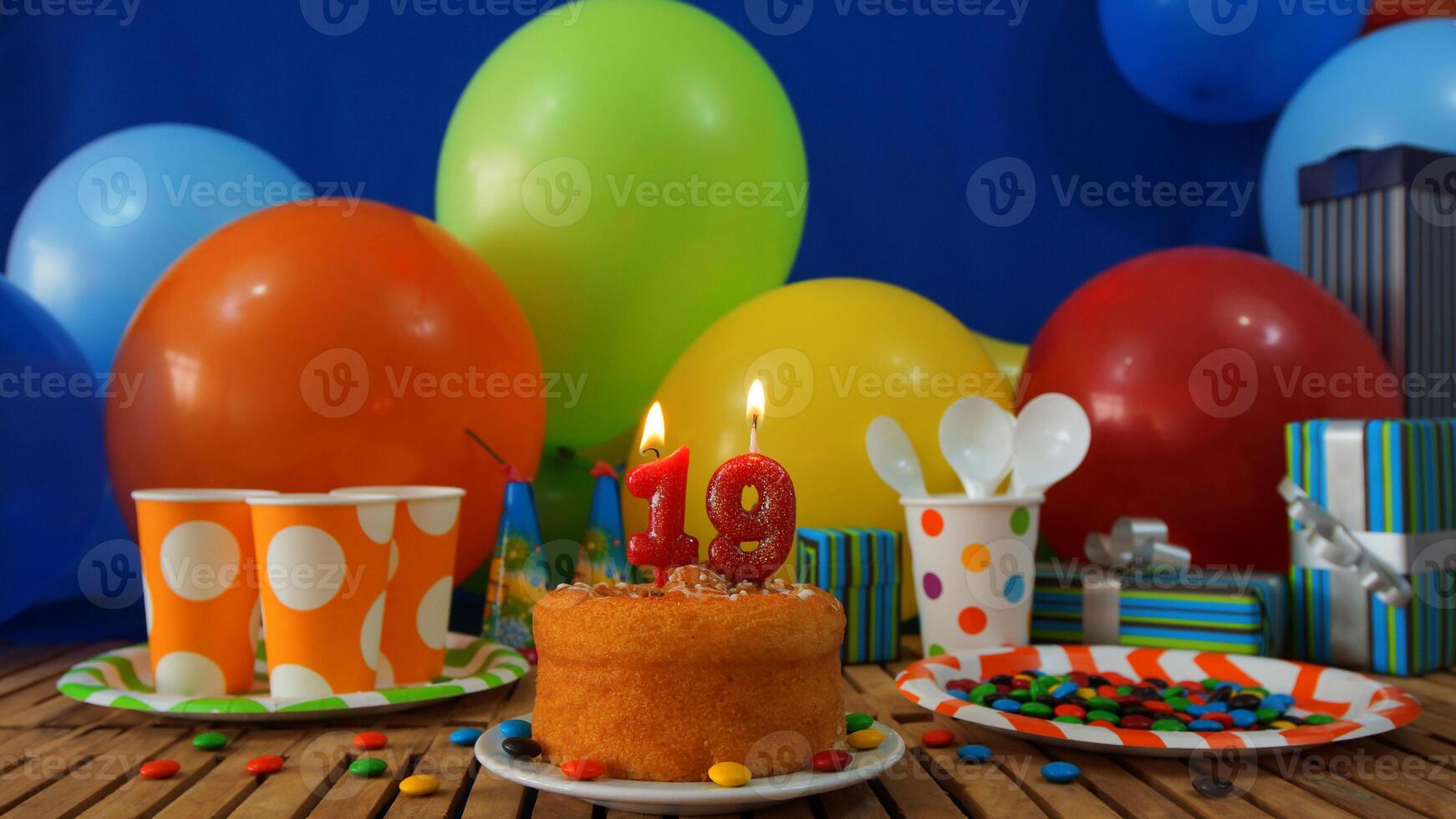 Birthday cake 19 on rustic wooden table with background of colorful balloons, gifts, plastic cups and plastic plate with candies and blue wall in the background photo
