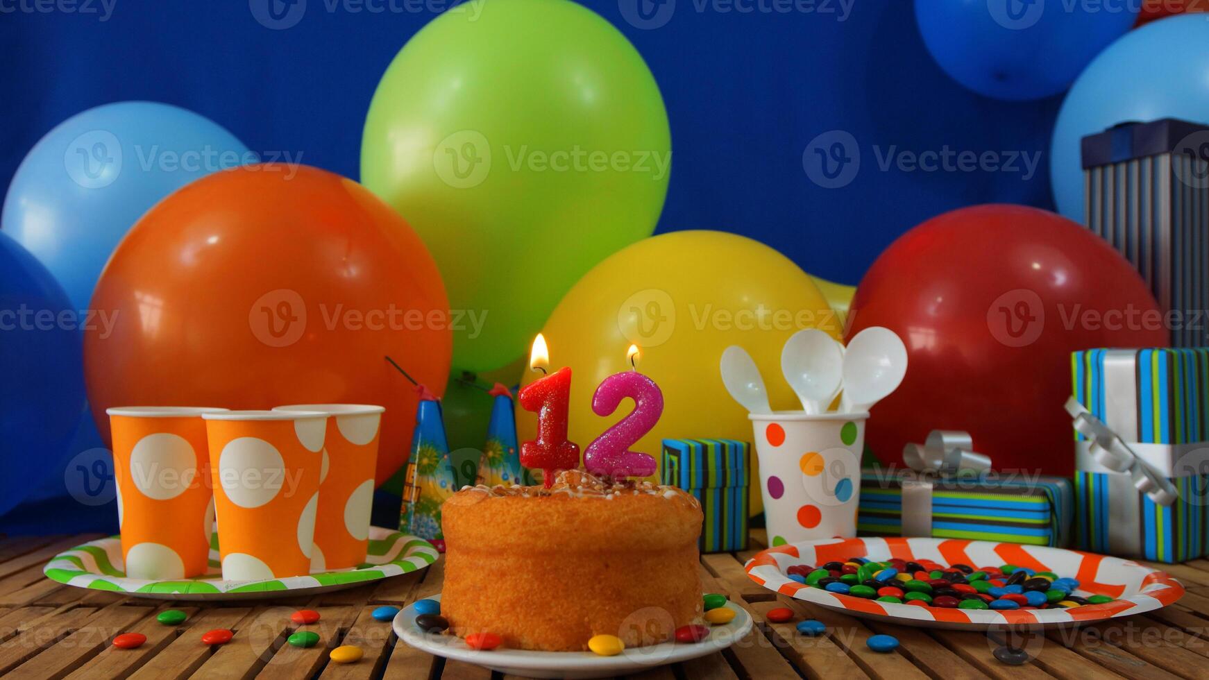 Birthday cake 12 on rustic wooden table with background of colorful balloons, gifts, plastic cups and plastic plate with candies and blue wall in the background photo
