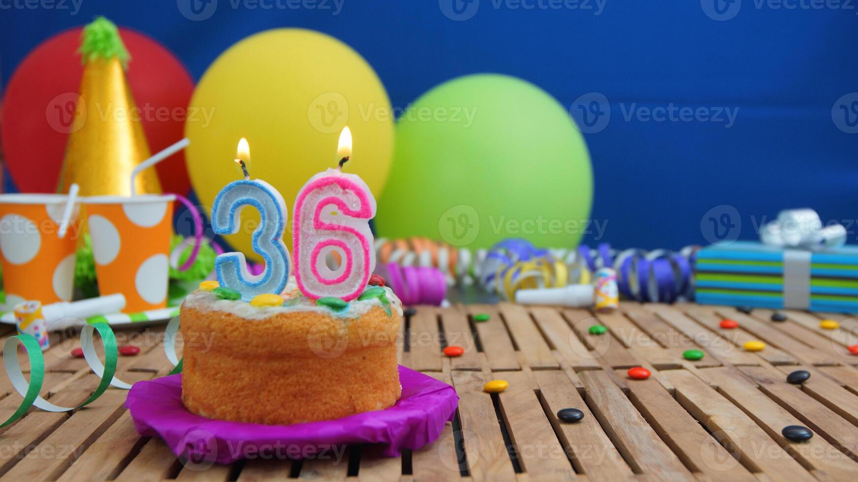 Birthday cake 36 with candles on rustic wooden table with background of colorful balloons, gifts, plastic cups and candies with blue wall in the background. Focus is on cake photo