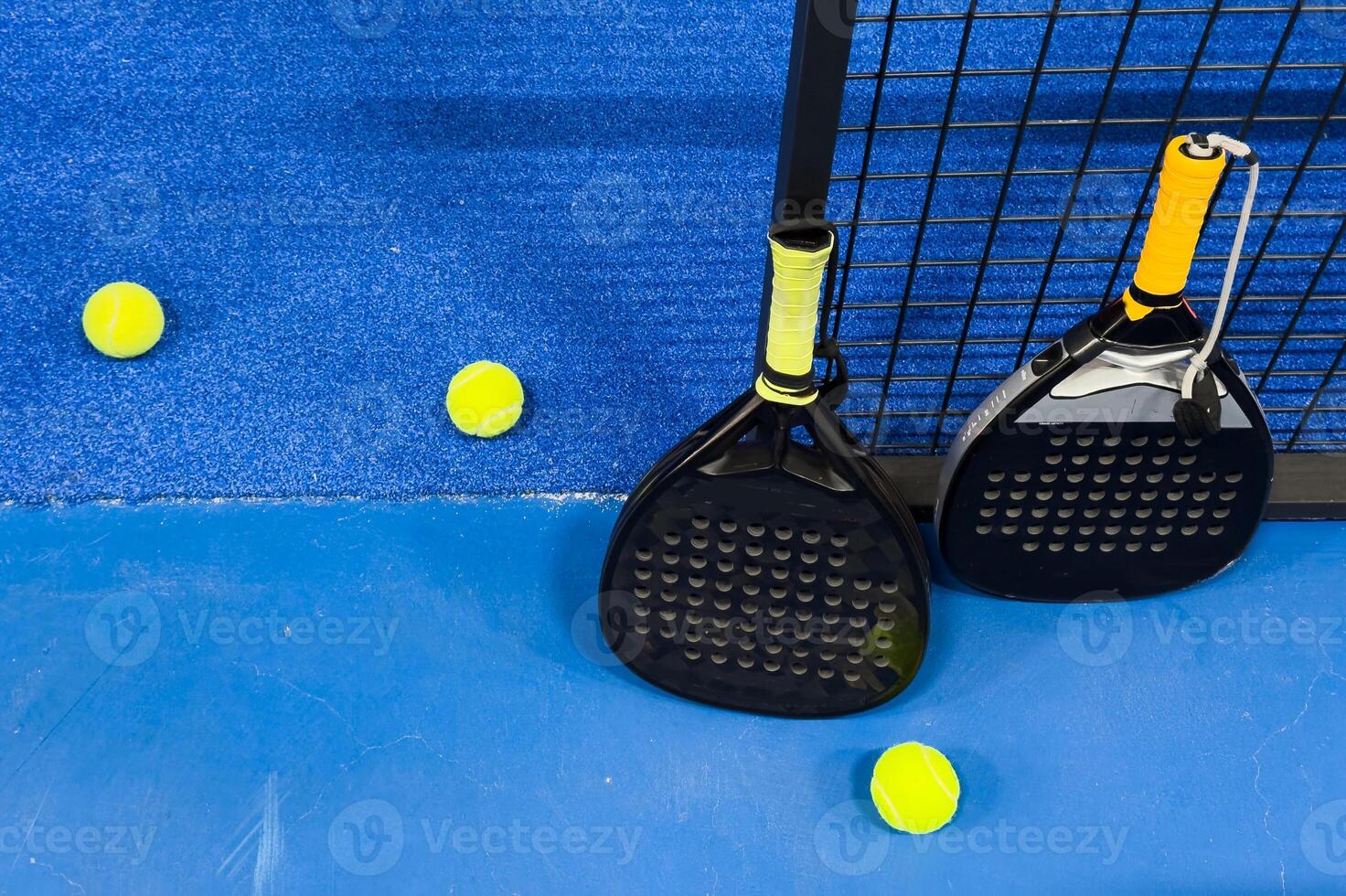 A padel or paddle tennis racket and a bright yellow tennis ball lie on a blue court. The net and a blurred figure of a player are visible in the background. photo