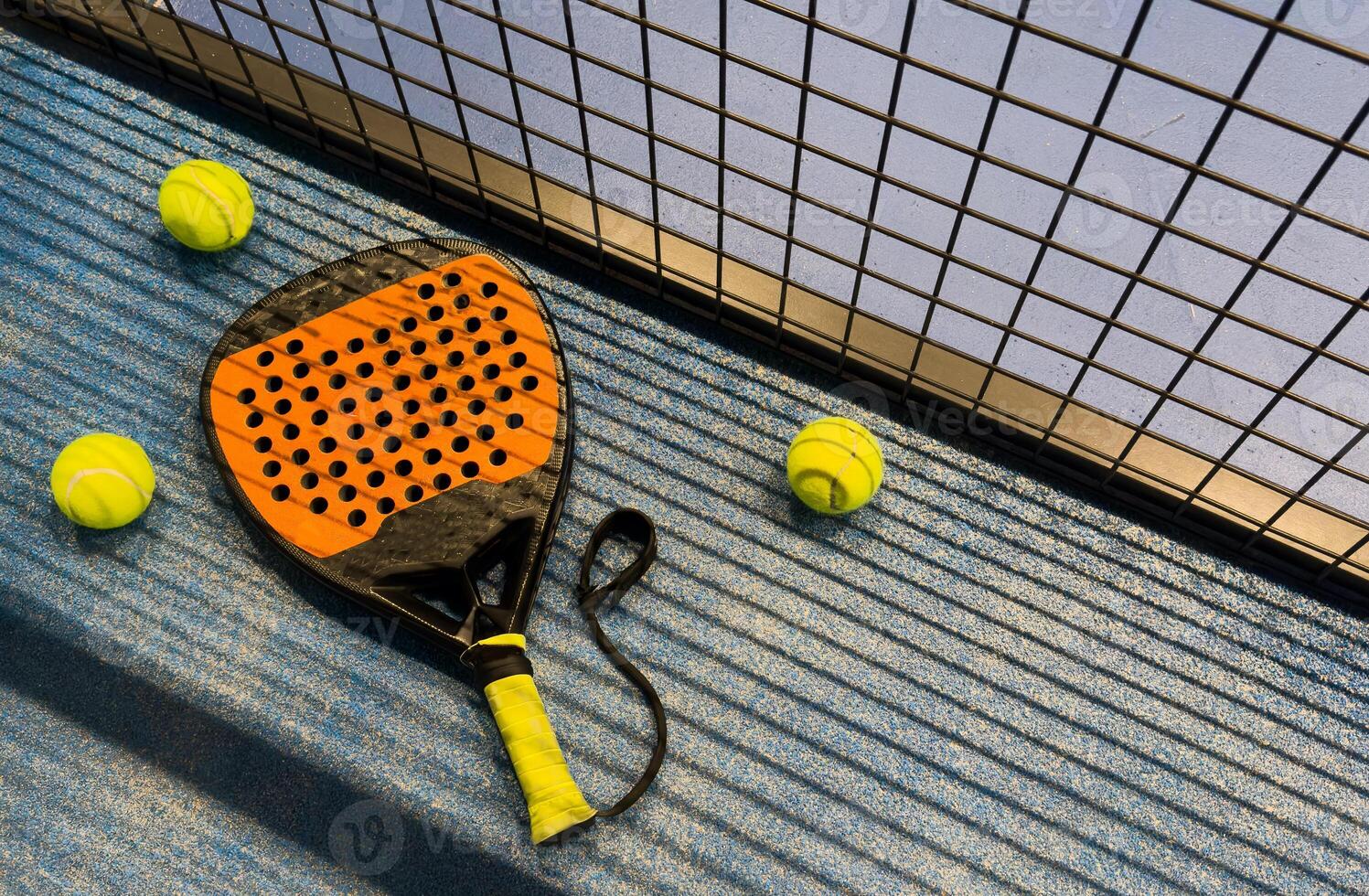 A padel or paddle tennis racket and a bright yellow tennis ball lie on a blue court. The net and a blurred figure of a player are visible in the background. photo