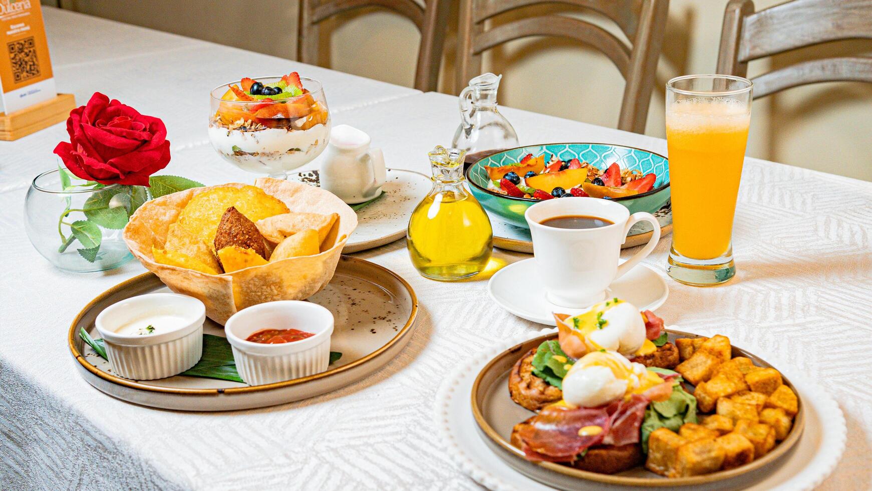 A breakfast table featuring coffee, orange juice, fruits, a gourmet plate with toast, burrata, serrano ham, tropical fruits, and traditional Caribbean fried snacks photo