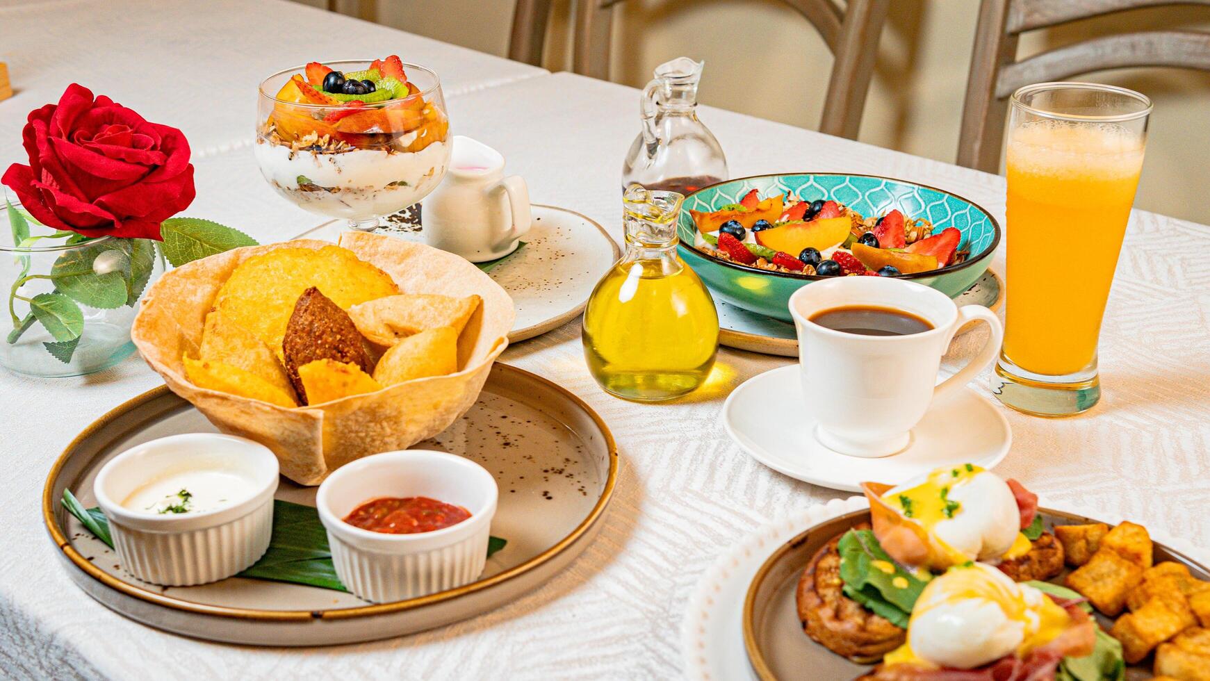 A breakfast table featuring coffee, orange juice, fruits, a gourmet plate with toast, burrata, serrano ham, tropical fruits, and traditional Caribbean fried snacks photo