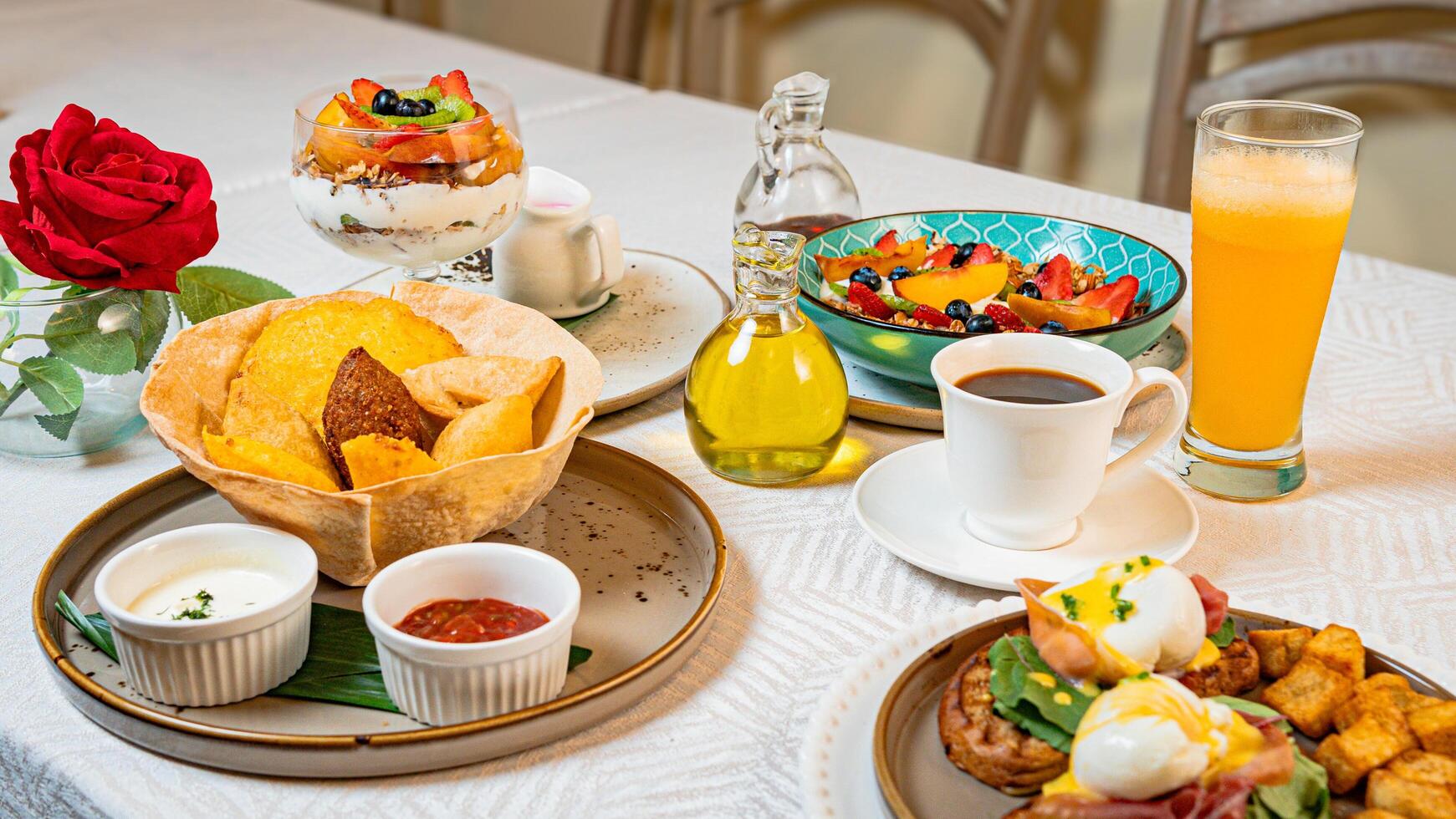 A breakfast table featuring coffee, orange juice, fruits, a gourmet plate with toast, burrata, serrano ham, tropical fruits, and traditional Caribbean fried snacks photo