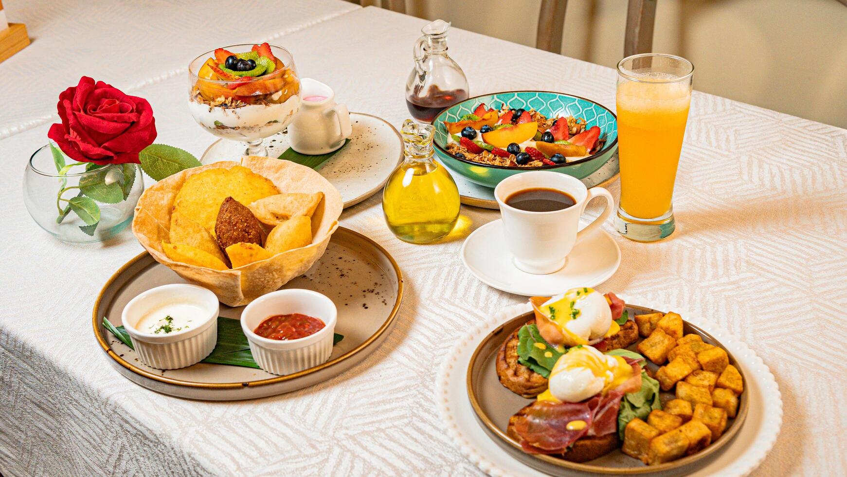 A breakfast table featuring coffee, orange juice, fruits, a gourmet plate with toast, burrata, serrano ham, tropical fruits, and traditional Caribbean fried snacks photo
