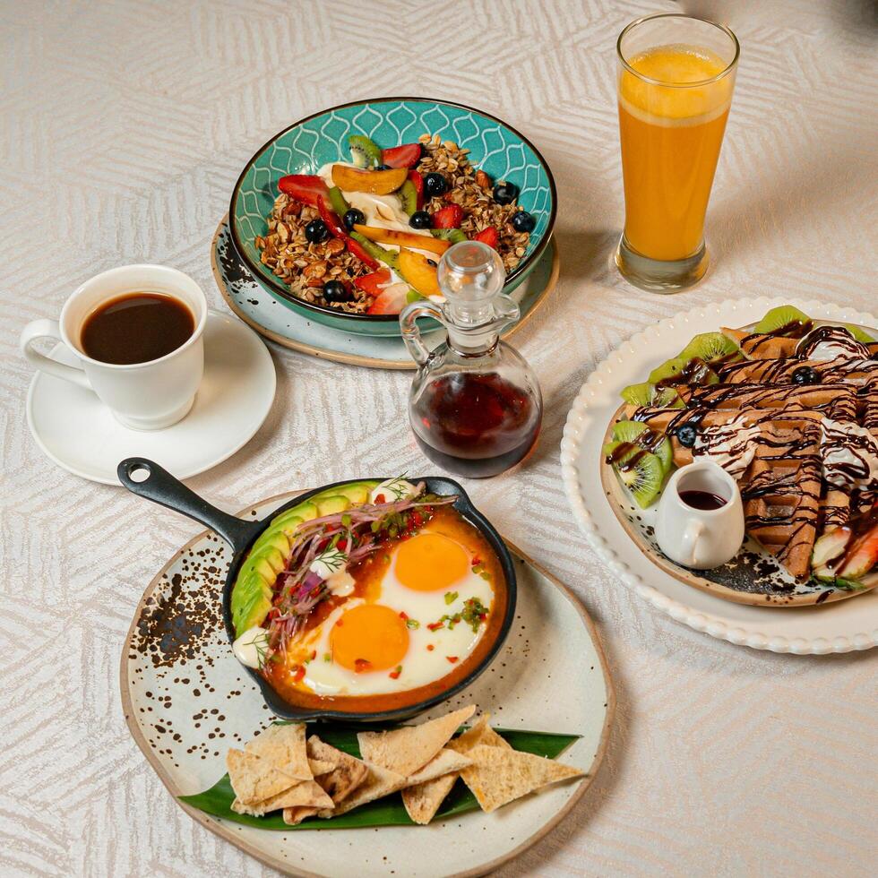 A breakfast table featuring coffee, orange juice, fruits, a gourmet plate with toast, burrata, serrano ham, tropical fruits, and traditional Caribbean fried snacks photo