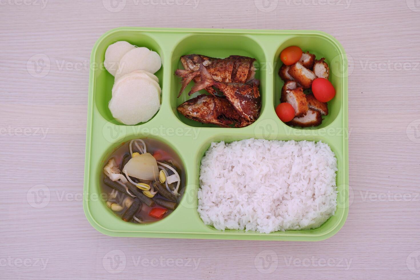A green tray with five compartments containing white rice, curry, sliced cucumbers and tomatoes, a boiled egg, quail eggs, fried chicken, and pieces of meat photo