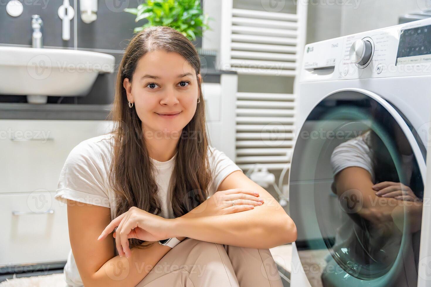 Young woman waiting for the washing machine to finish, looking at the camera next to washing machine door. High quality photo