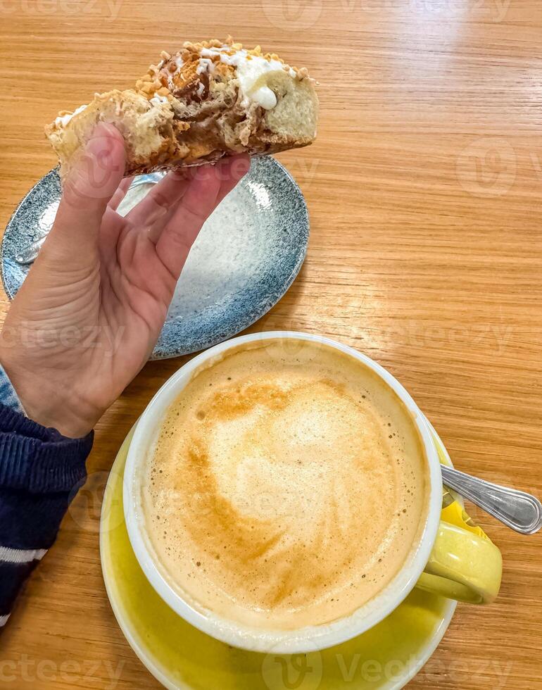 female hand holding bitten bun and yellow cup of coffee on wooden table . High quality photo
