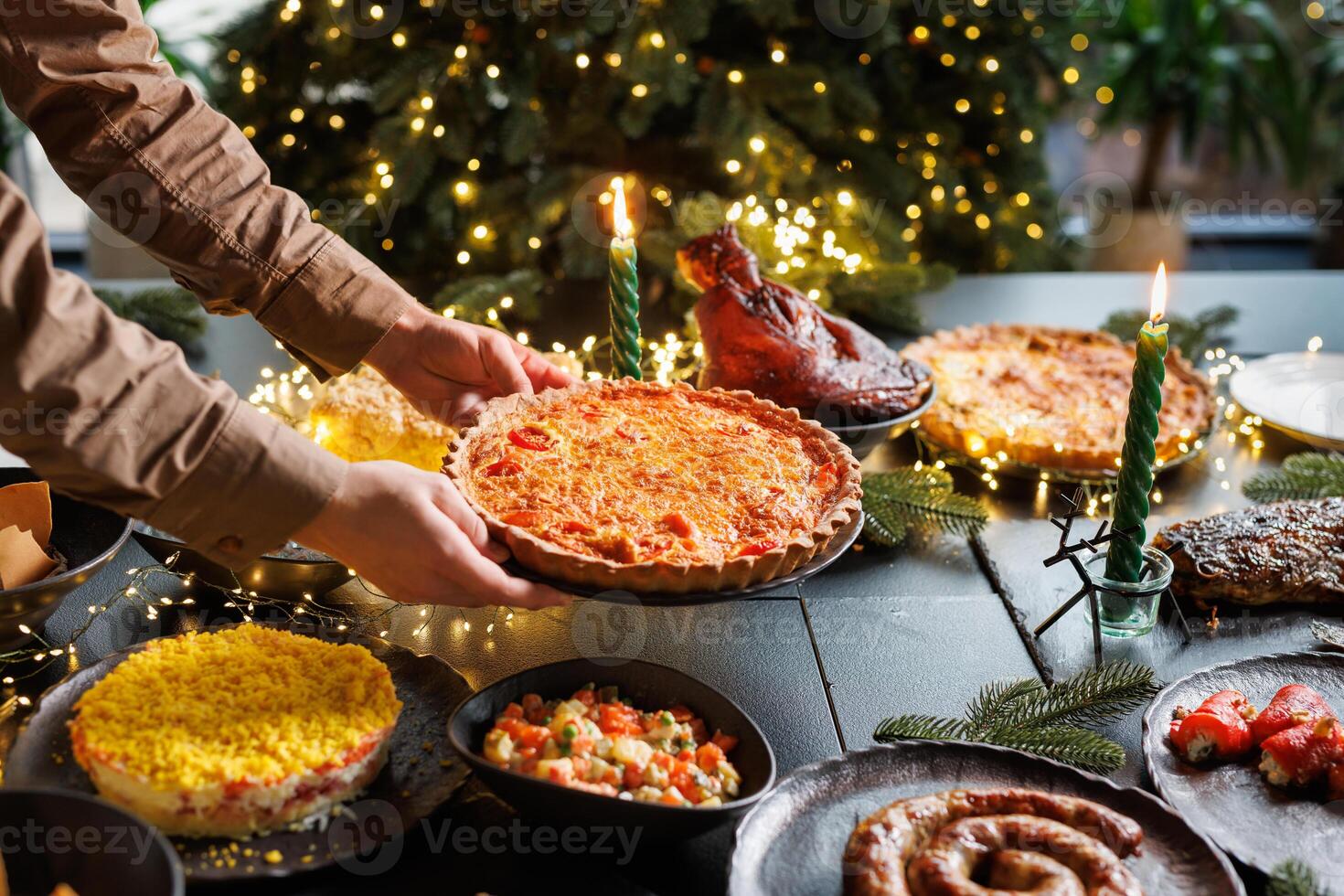 Man places Christmas quiche pie on festive table surrounded by many dishes in cozy evening atmosphere with Christmas tree and garlands photo