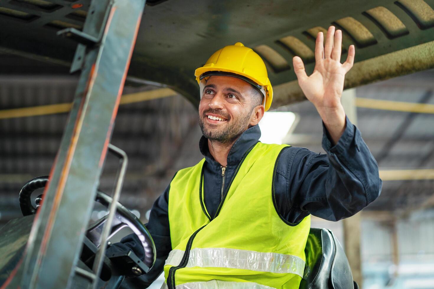 man in a yellow vest and a hard hat is waving from the cab of a forklift photo
