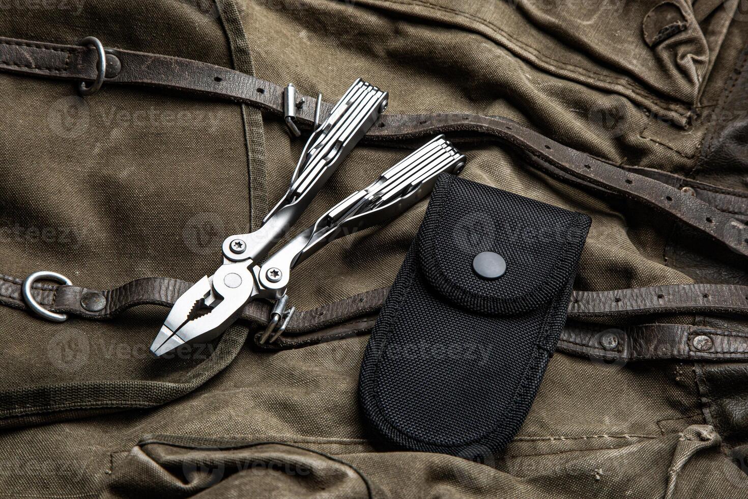 A multi-tool with pliers is placed on a brown canvas bag with leather straps, accompanied by a black nylon sheath, showcasing its utility and rugged design photo