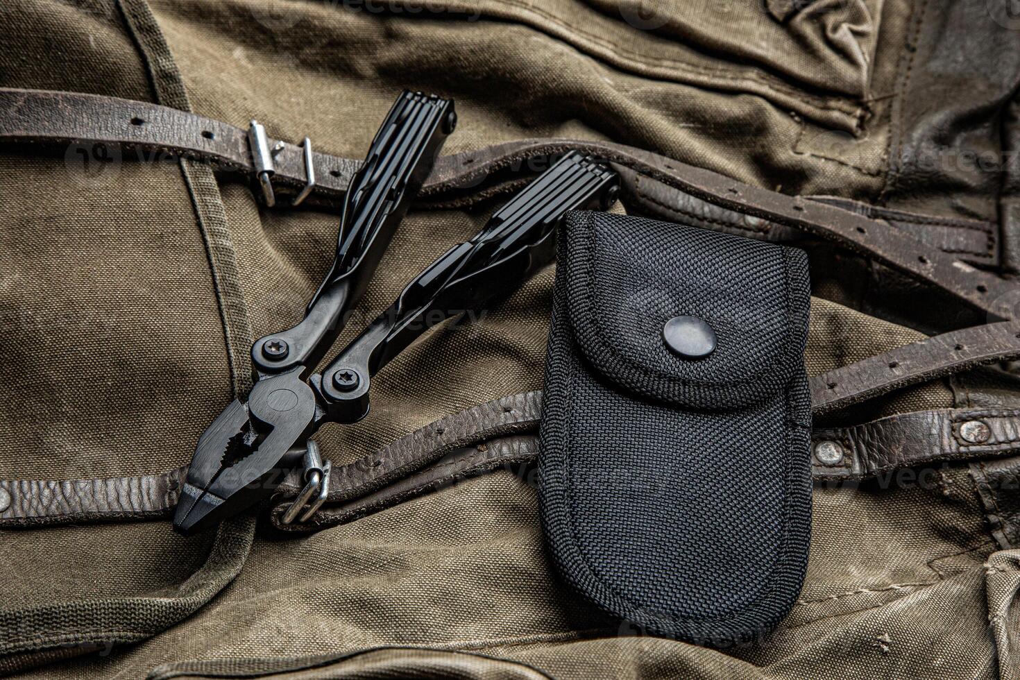 A multi-tool with pliers is placed on a brown canvas bag with leather straps, accompanied by a black nylon sheath, showcasing its utility and rugged design photo