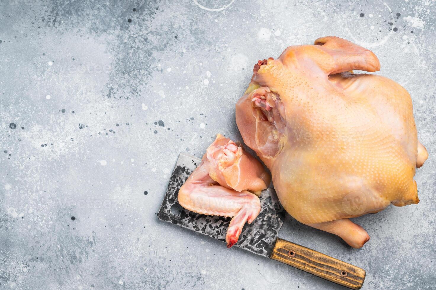 A whole raw chicken and a single chicken wing lay on a gray countertop with a cleaver photo