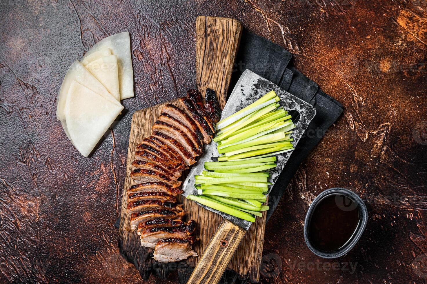 Sliced Peking Duck served with cucumber, green onion, and wheaten chinese pancakes on a meat cleaver. Dark background. Top view photo
