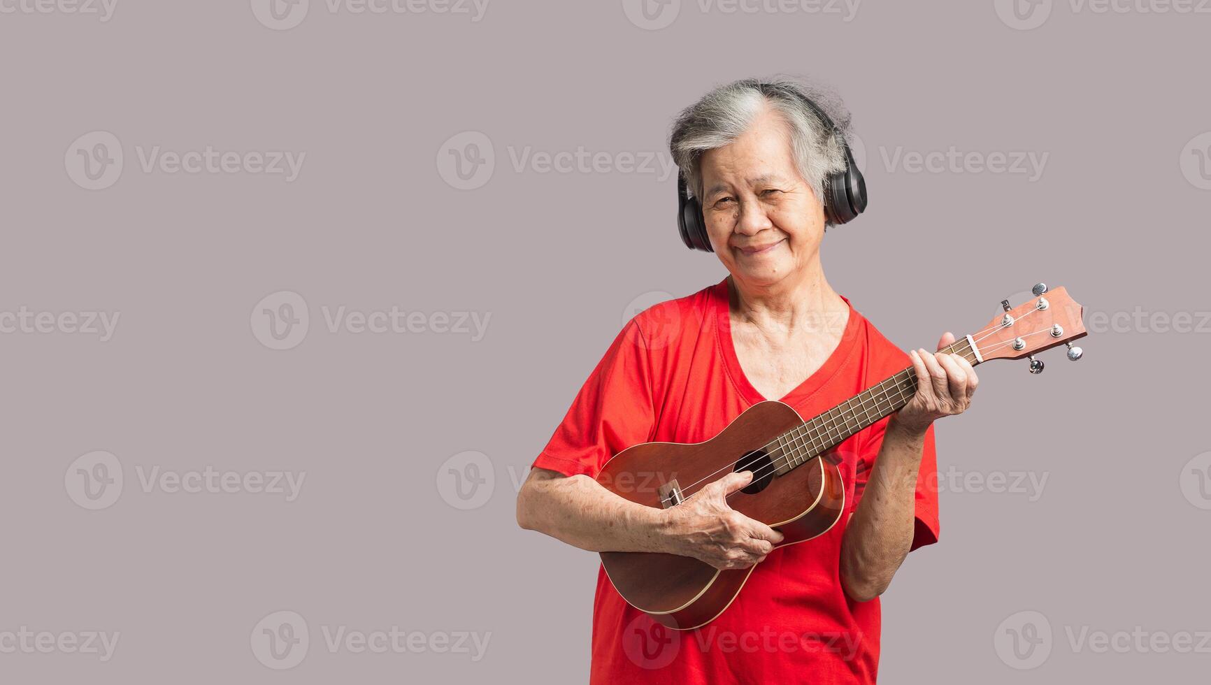 Portrait of an elderly Asian woman with short gray hair playing the ukulele with a smile while standing on a gray background photo