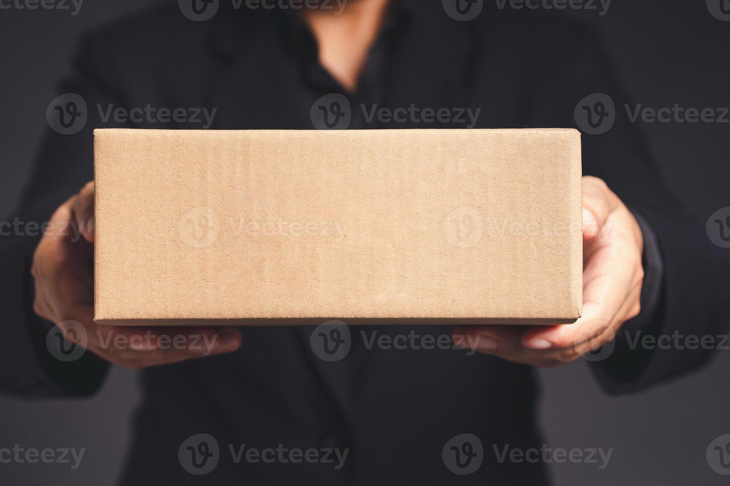 Businessman in a black suit holds and points to a cardboard box while standing on a gray background photo