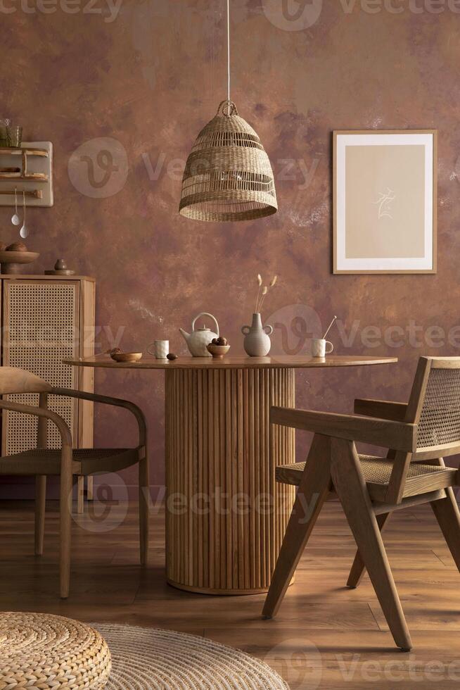 Stylish boho vintage dining room interior with poster mock up. Round table with rattan armchair, round carpet with pouf, vase with dried flowers and cup. Brown wall and rattan lamp. Mock up poster. photo