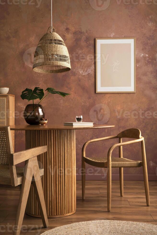 Stylish boho vintage dining room interior with poster mock up. Round table with rattan armchair, vase with dried flowers and cup. Brown wall and rattan lamp. Mock up poster. Template. photo
