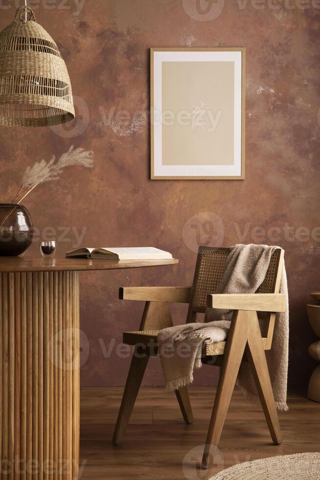 Stylish boho vintage dining room interior with poster mock up. Round table with rattan armchair, vase with dried flowers and cup. Brown wall and rattan lamp. Mock up poster. Template. photo
