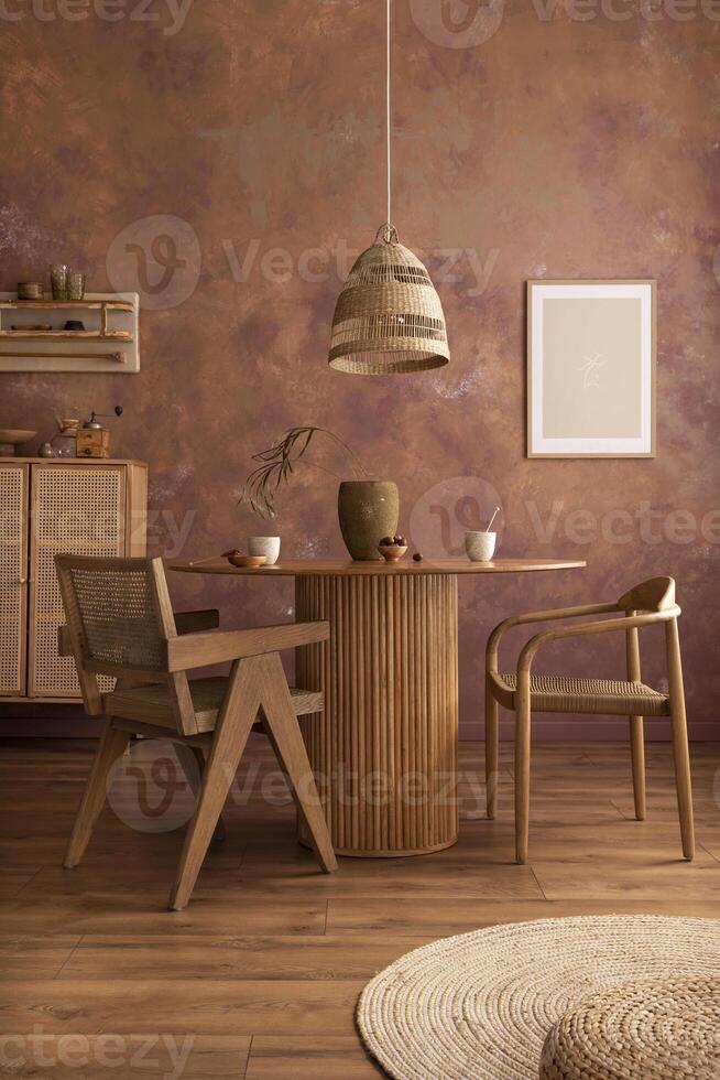 Stylish boho vintage dining room interior with poster mock up. Round table with rattan armchair, vase with dried flowers and cup. Brown wall and rattan lamp. Mock up poster. Template. photo